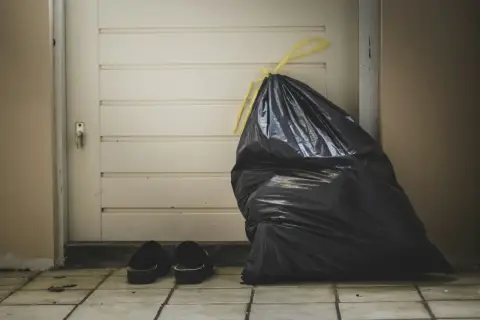 A full black trash bag and a pair of slippers sit by a closed door, symbolizing the need for lean budgeting to discard unnecessary expenses.