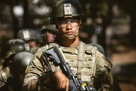 A young soldier, determined and focused, stands ready during far bootcamp. He wears a helmet, protective eyewear, and camouflage fatigues, holding his rifle.