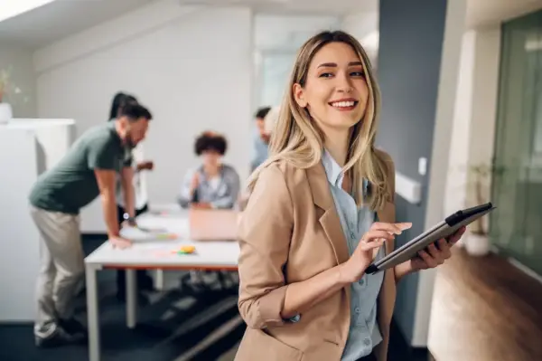 A smiling woman with a tablet, likely pursuing her masters in leadership and management, stands in an office as colleagues collaborate in the background.